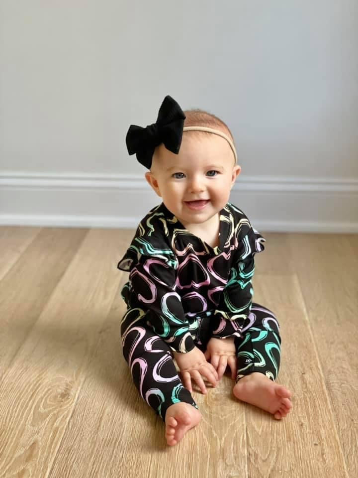 Baby sitting on a wooden floor wearing a pink and green onesie.